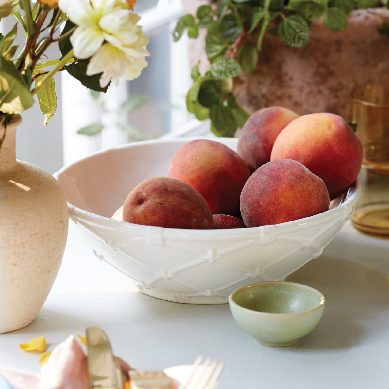 White bowl filled with ripe peaches on a table beside flowers and a small green dish.