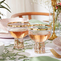 Two gold-rimmed glasses with a floral tablecloth and decorative elements in the background.